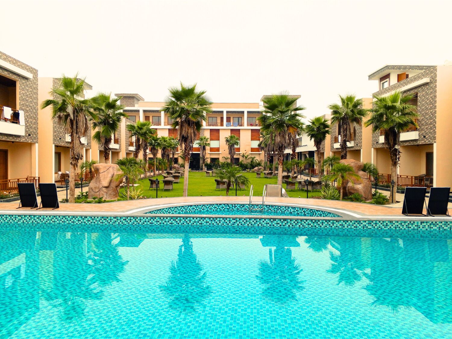 A resort pool area with a clear blue pool, lounge chairs, palm trees, and a building with balconies.