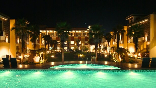 A nighttime view of a hotel with a swimming pool, palm trees, and illuminated buildings. - The Maasai Mara Resort Water and Adventure Park Corbett (Ramnagar)