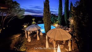 Vista nocturna de una villa con piscina, rodeada de altos cipreses y sombrillas exteriores.