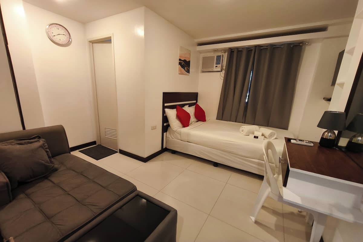Een kleine hotelkamer met een bed, een slaapbank, een tafeltje, een stoel, een klok, een airconditioner en een raam met gordijnen.