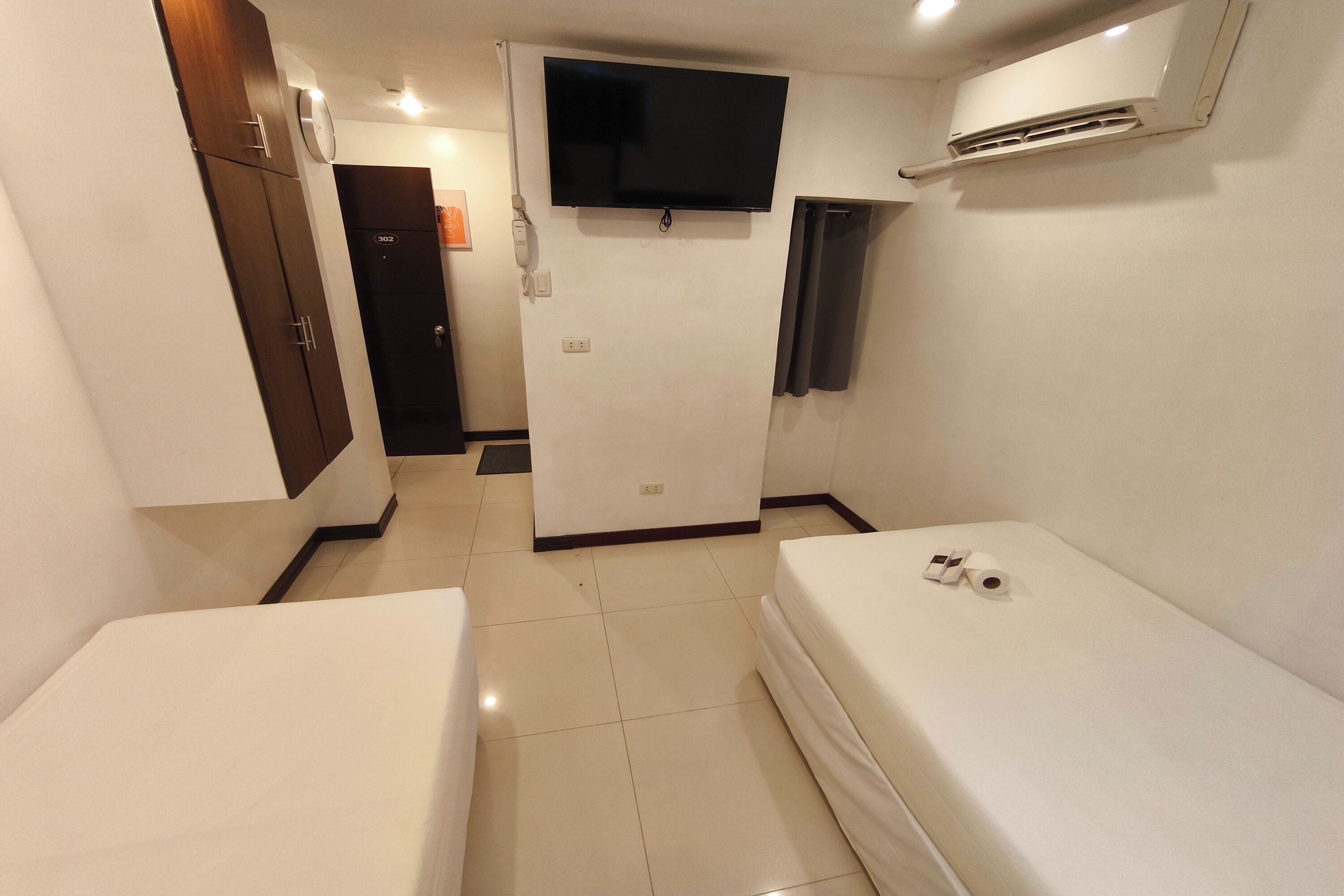 Habitación de hotel con dos camas individuales, un televisor montado en la pared, un equipo de aire acondicionado y una puerta que da a otra habitación.
