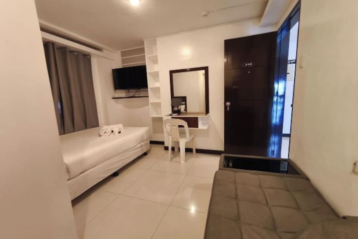 A hotel room with a bed, a desk, a chair, a TV, and a door with a number.