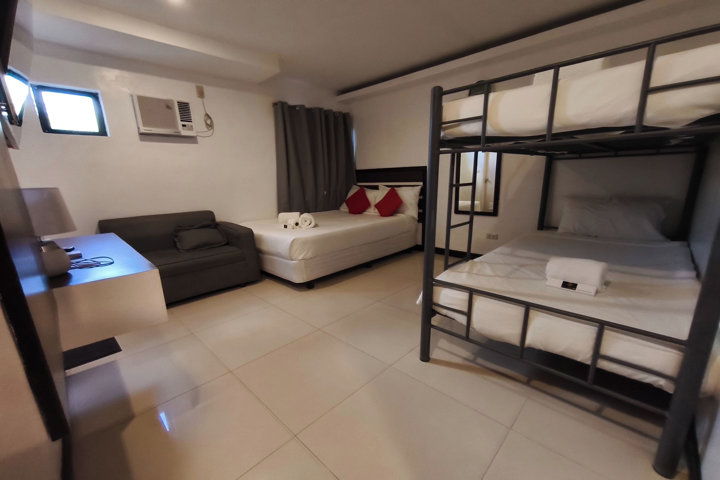 Habitación compacta de hotel con litera, cama individual, escritorio con silla, sofá y ventana con cortinas.
