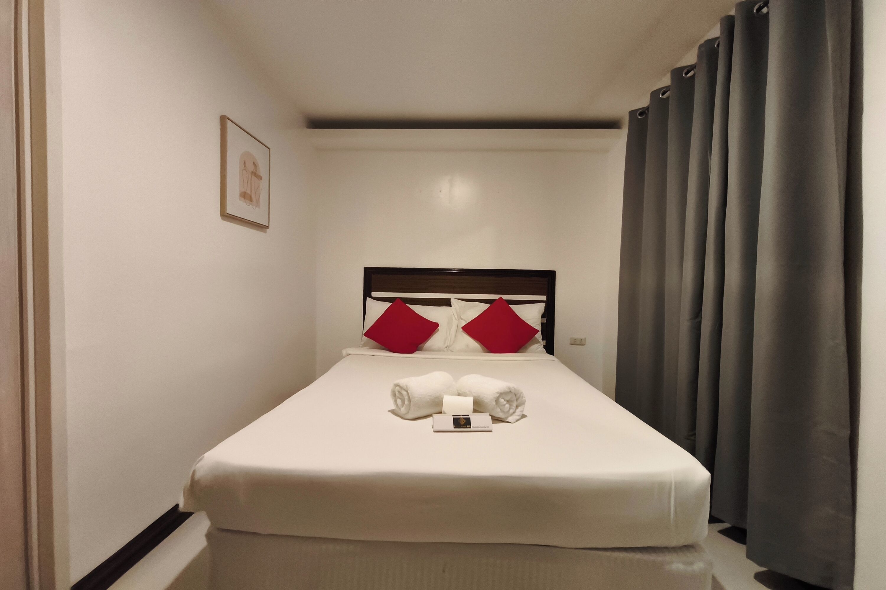 A narrow hotel room with a single bed, red pillows, and a small framed picture on the wall.