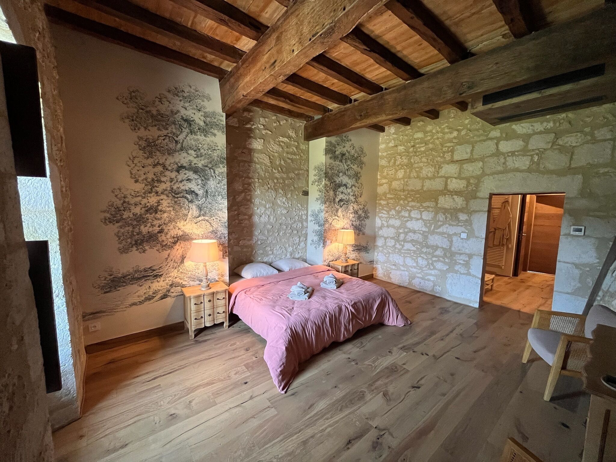 A bedroom with a stone wall, wooden beams, a bed with a pink bedspread, two bedside tables with lamps, and a wooden chair.