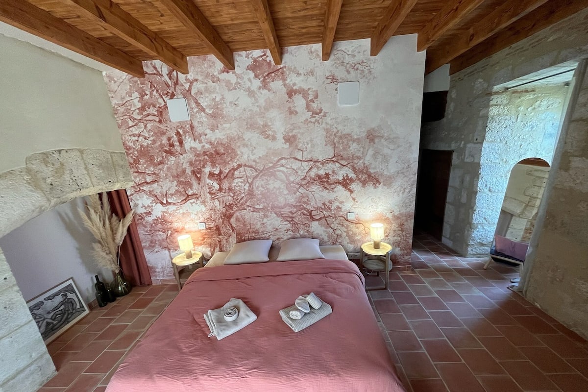 A bedroom with a large bed, two bedside lamps, a wall mural, and a rustic stone wall.