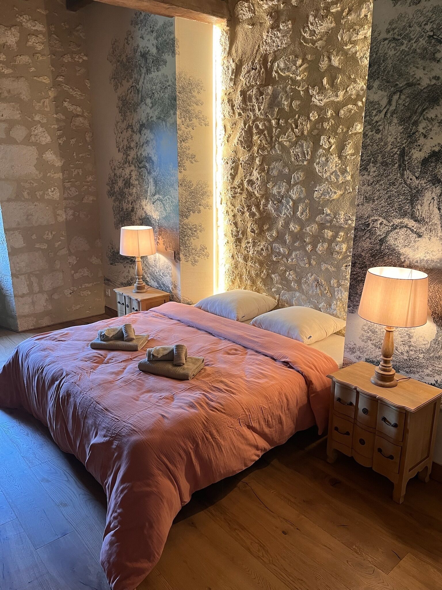 A bedroom with a large bed, two bedside tables with lamps, a wooden floor, and textured walls.