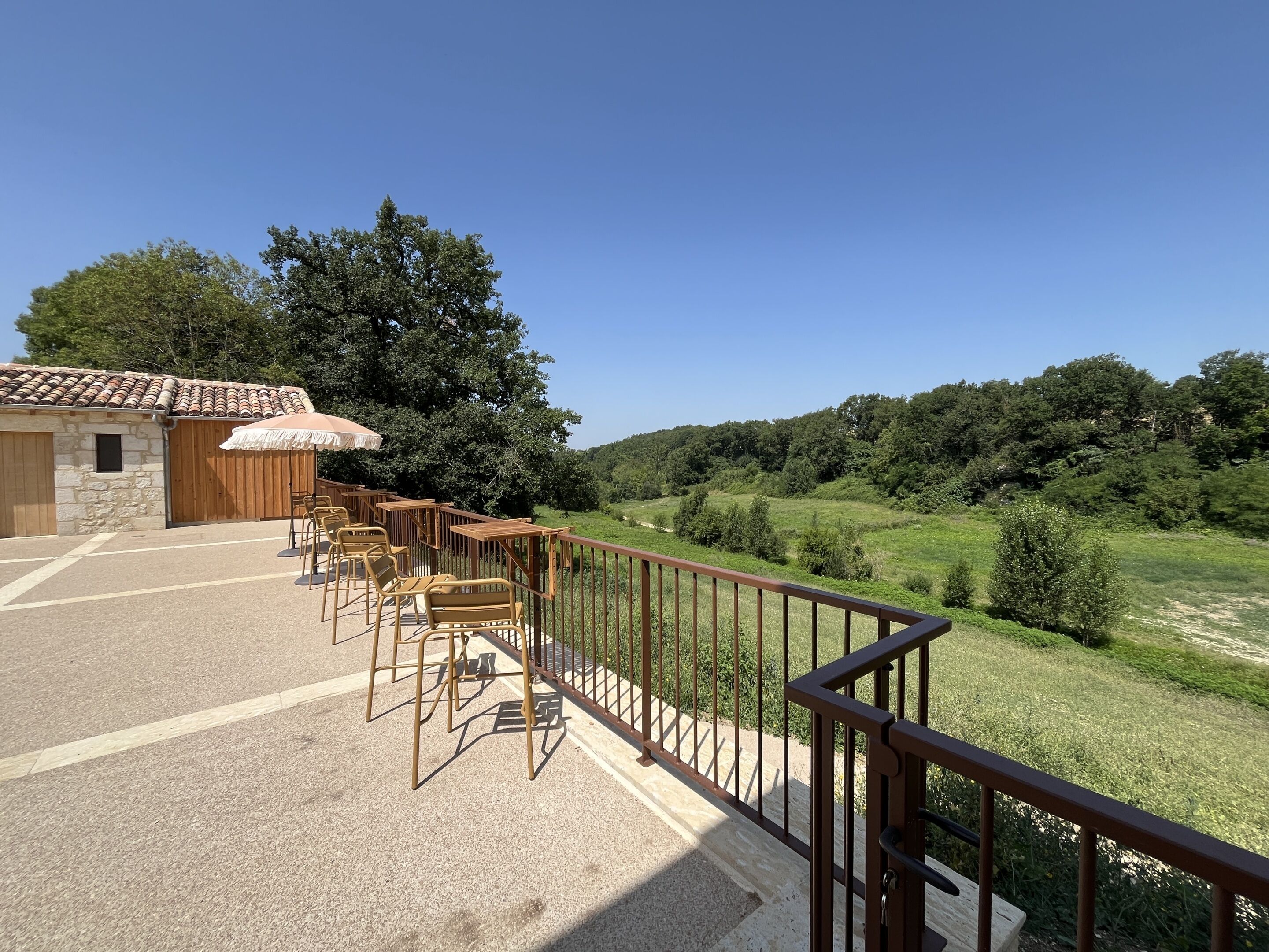 An outdoor seating area with wooden tables and chairs, a stone building with a tiled roof, and a view of a green hillside.