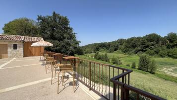 An outdoor seating area with wooden tables and chairs, a stone building with a tiled roof, and a view of a green hillside.