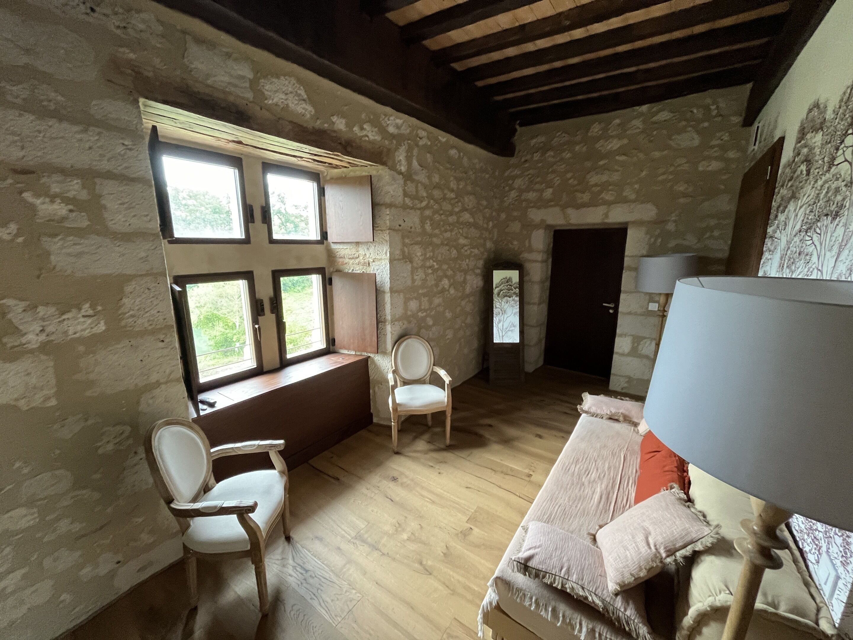 Una habitación con paredes de piedra, vigas de madera, un ventanal con múltiples cristales, dos sillas blancas, un banco de madera, una lámpara y una puerta.