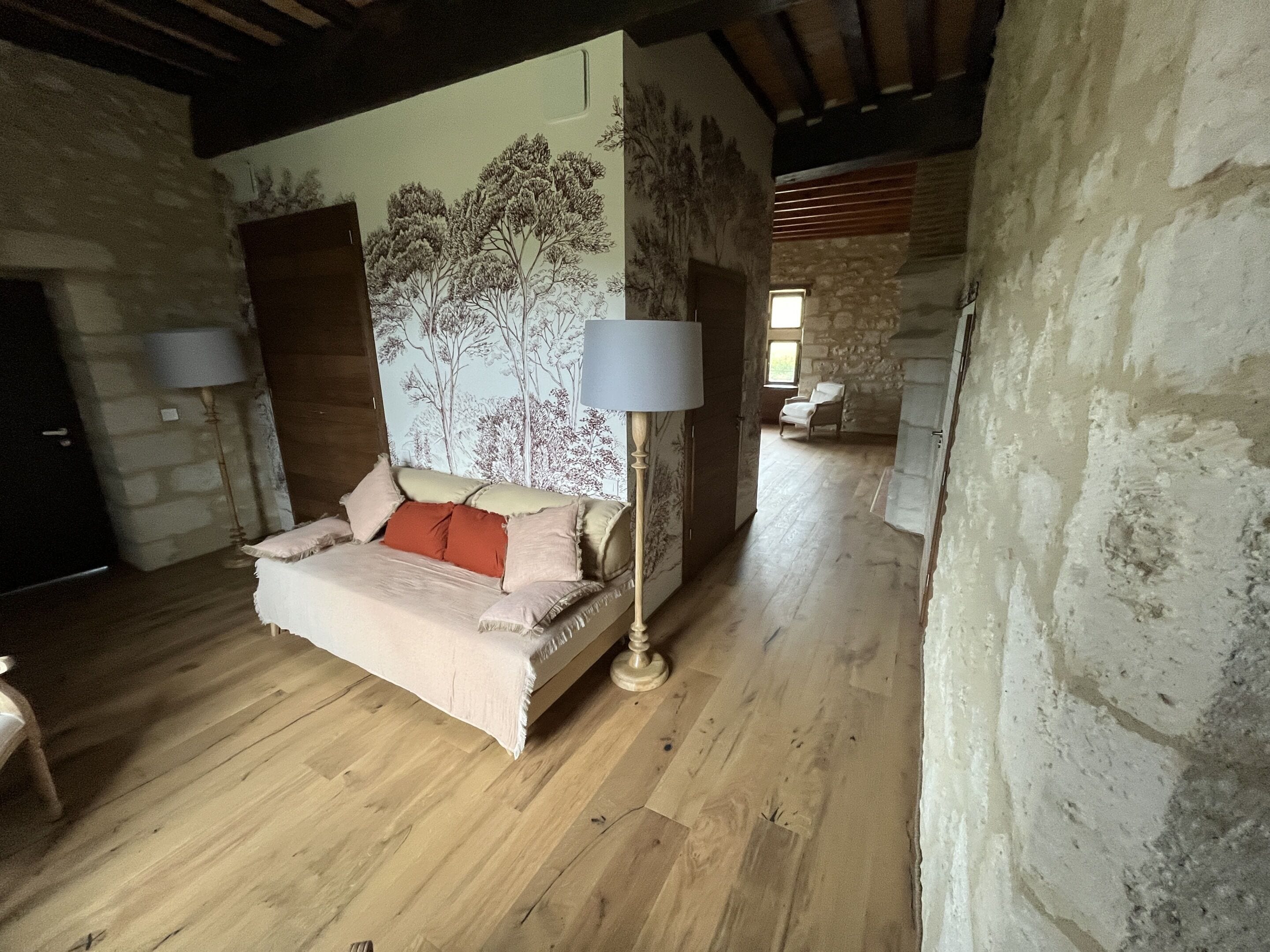 Ein gemütliches Zimmer mit Holzboden, einem Bett mit beiger und roter Bettdecke und einer Wandmalerei von Bäumen.
