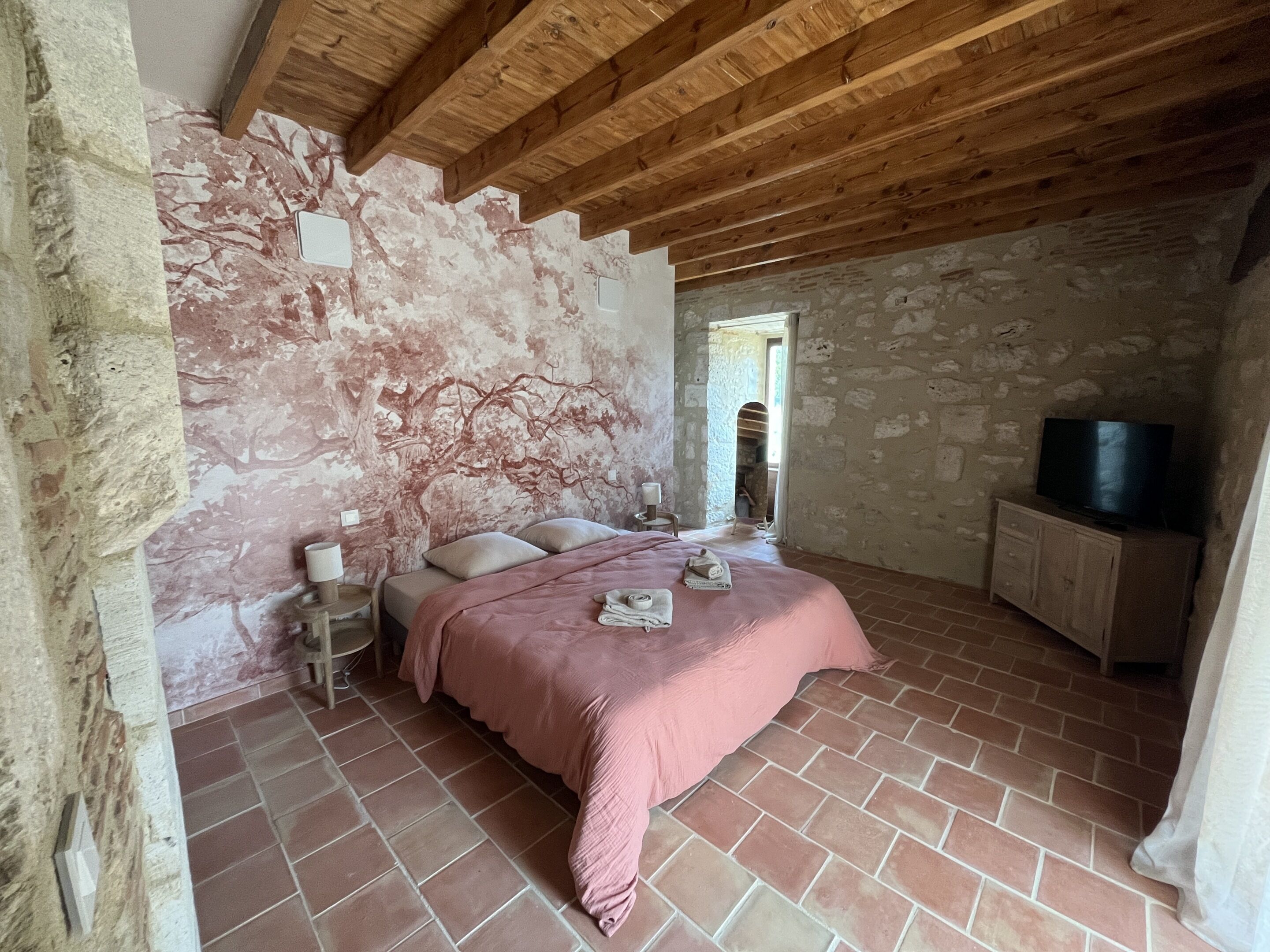 A bedroom with a stone wall, a wooden ceiling, a bed with a pink blanket, a bedside table with a lamp, and a TV on a wooden cabinet.