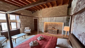 Una habitación con chimenea de piedra, cama con ropa de cama roja, un sillón blanco y techo de madera.