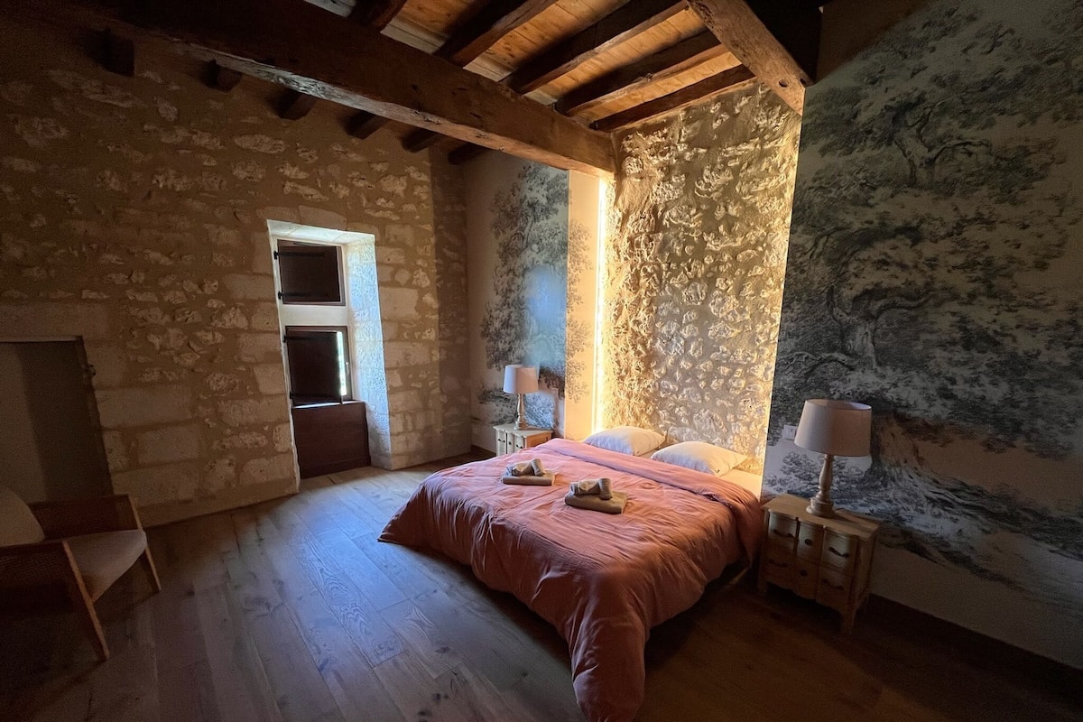 A bedroom with a stone wall, wooden beams, a bed with a pink bedspread, two bedside lamps, and a small table with a lamp.
