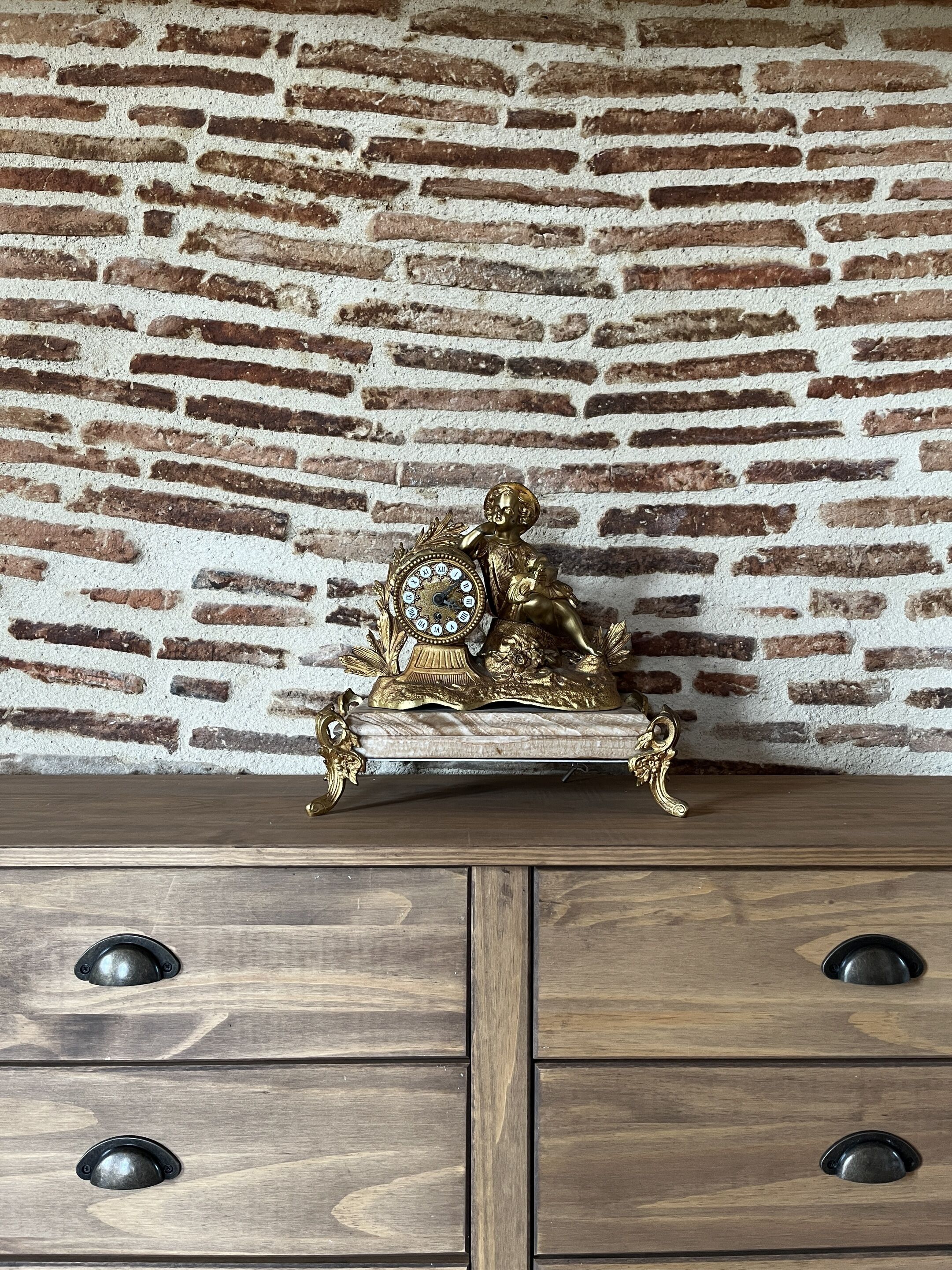 A wooden dresser with three drawers and a decorative brass clock with a cherub figure.