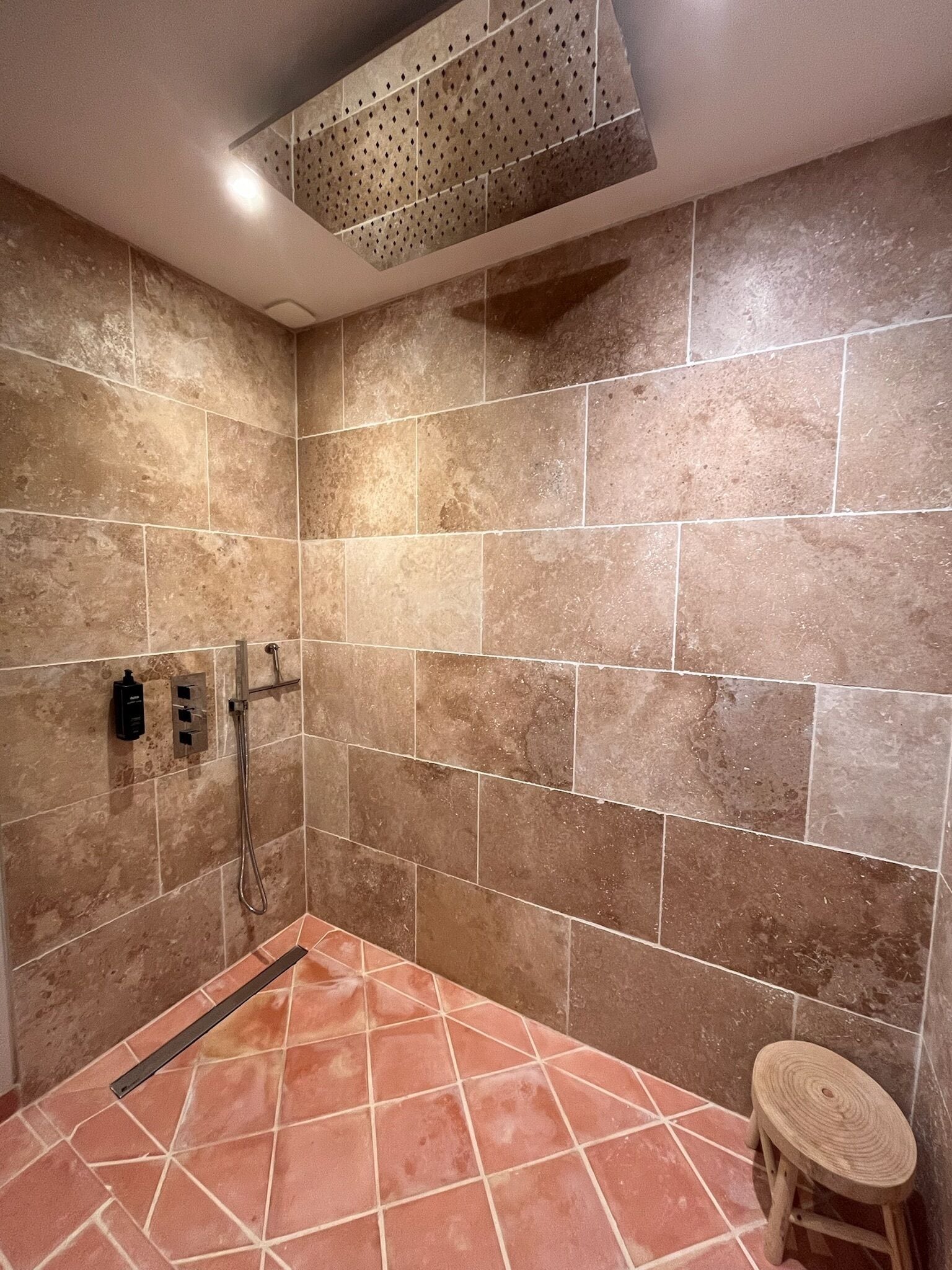 Un área de ducha con azulejos color beige, un cabezal de ducha de mano y un taburete pequeño.