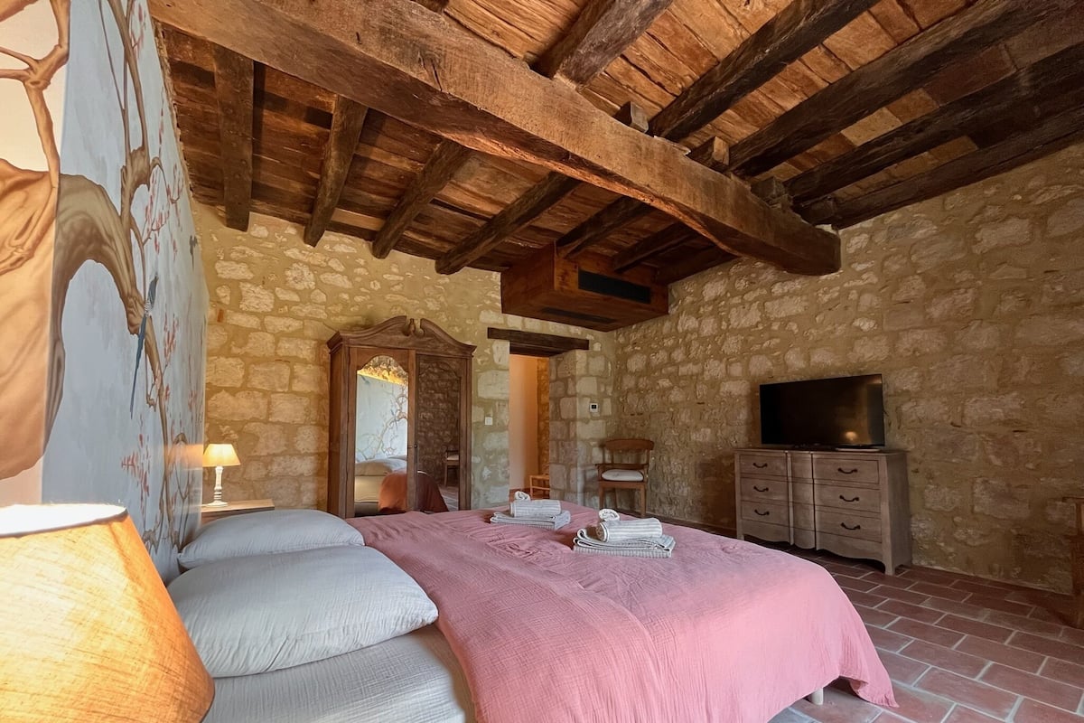 A bedroom with a stone wall, wooden beams, a bed with a pink blanket, a TV on a dresser, and a lamp.