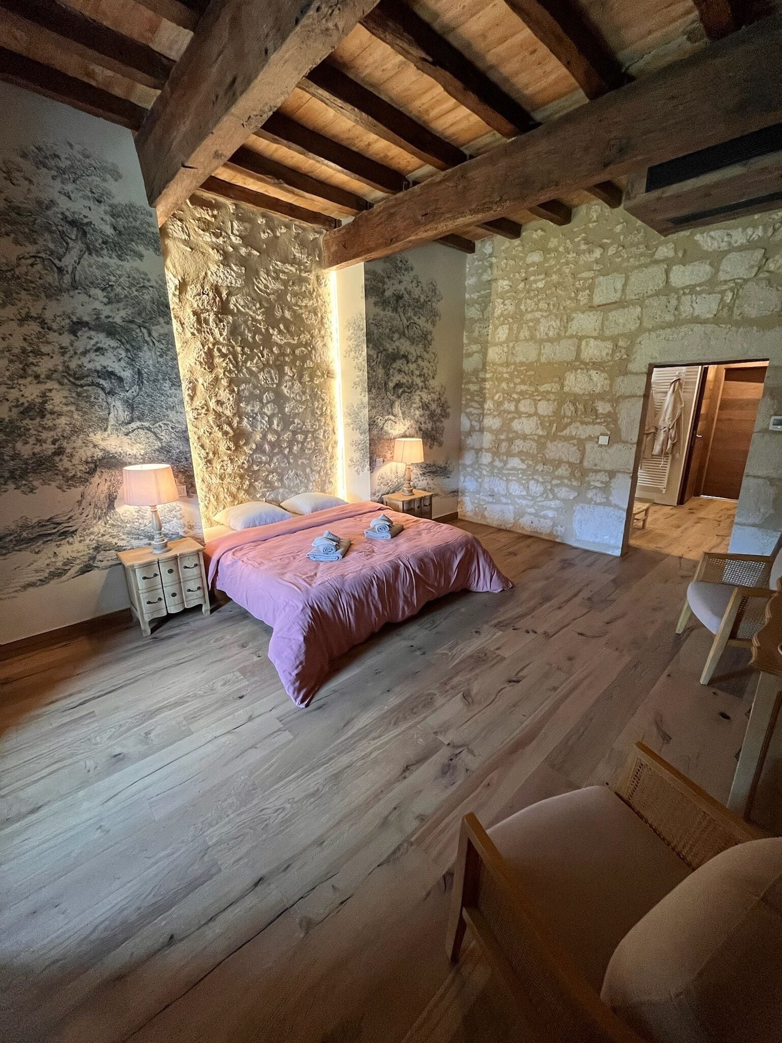 Une chambre avec un mur en pierre, des poutres apparentes, un lit recouvert d’une couvre-lit rose, deux tables de chevet avec des lampes et une armoire en bois.