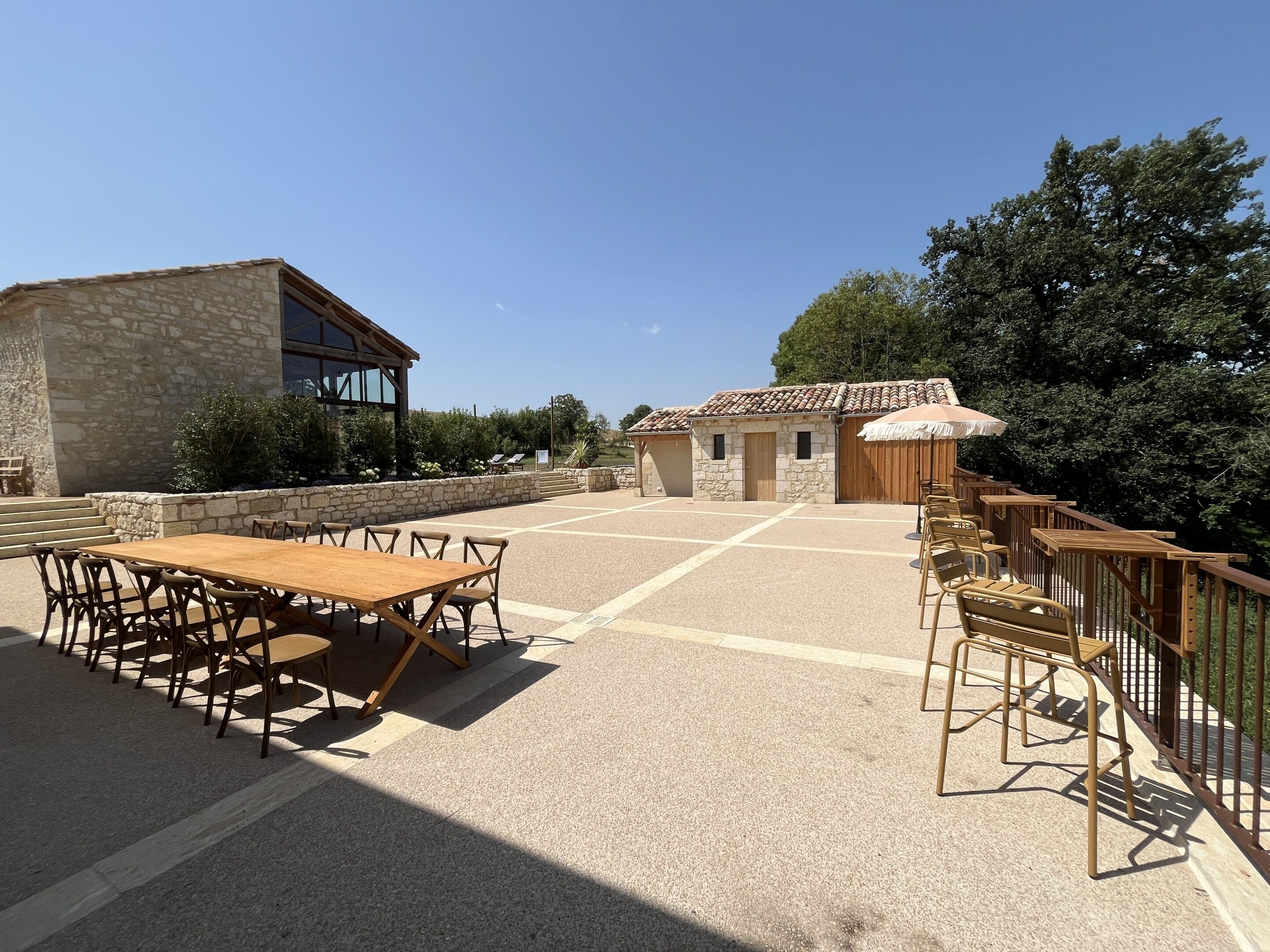 A spacious outdoor patio with a wooden table and chairs, stone walls, and a wooden building with a tiled roof.
