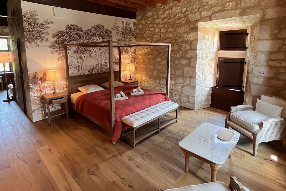 A cozy bedroom with a four-poster bed, a stone wall, and a wooden ceiling.