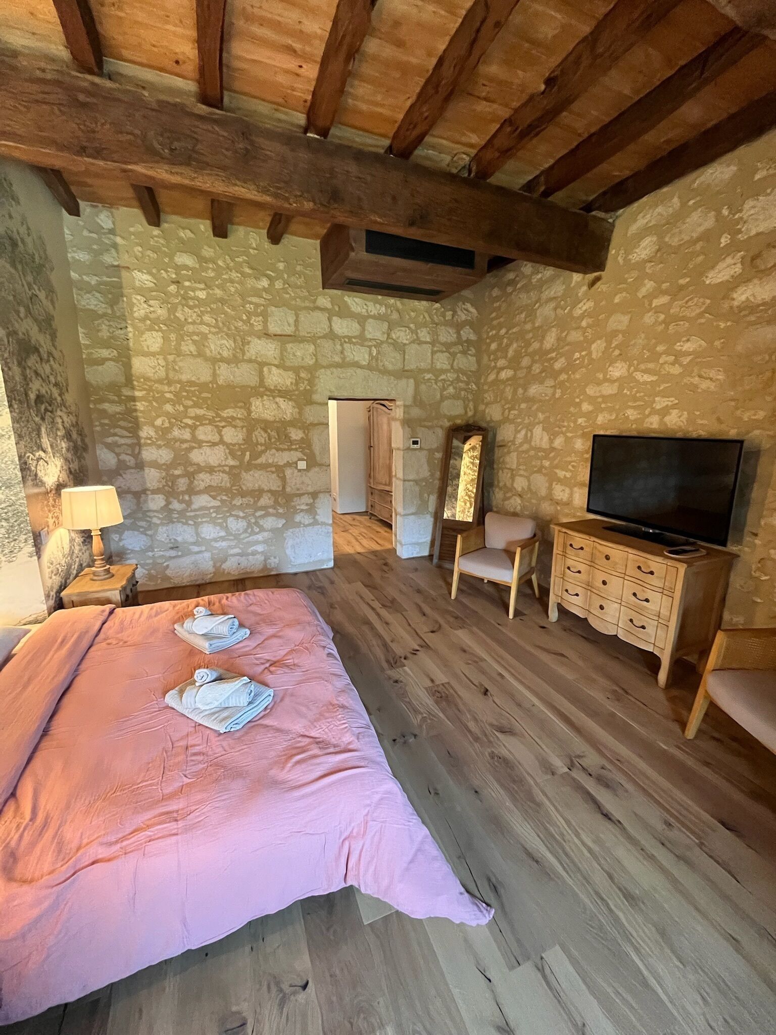 A bedroom with a stone wall, wooden floor, a bed with pink bedding, a TV, a dresser, and a mirror.