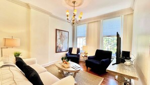 A living room with a sofa, armchairs, a coffee table, a chandelier, and large windows. - HEART of Old Town! Historic 1BR! 99 Walk Score! (Alexandria)