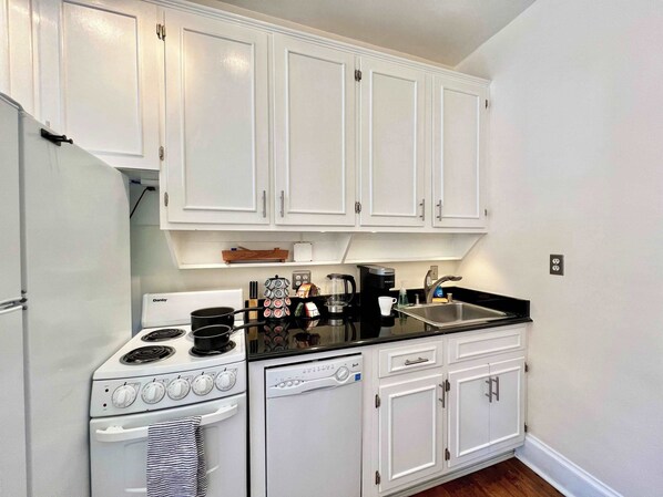 Una cocina compacta con armarios blancos, estufa, lavavajillas y cafetera.