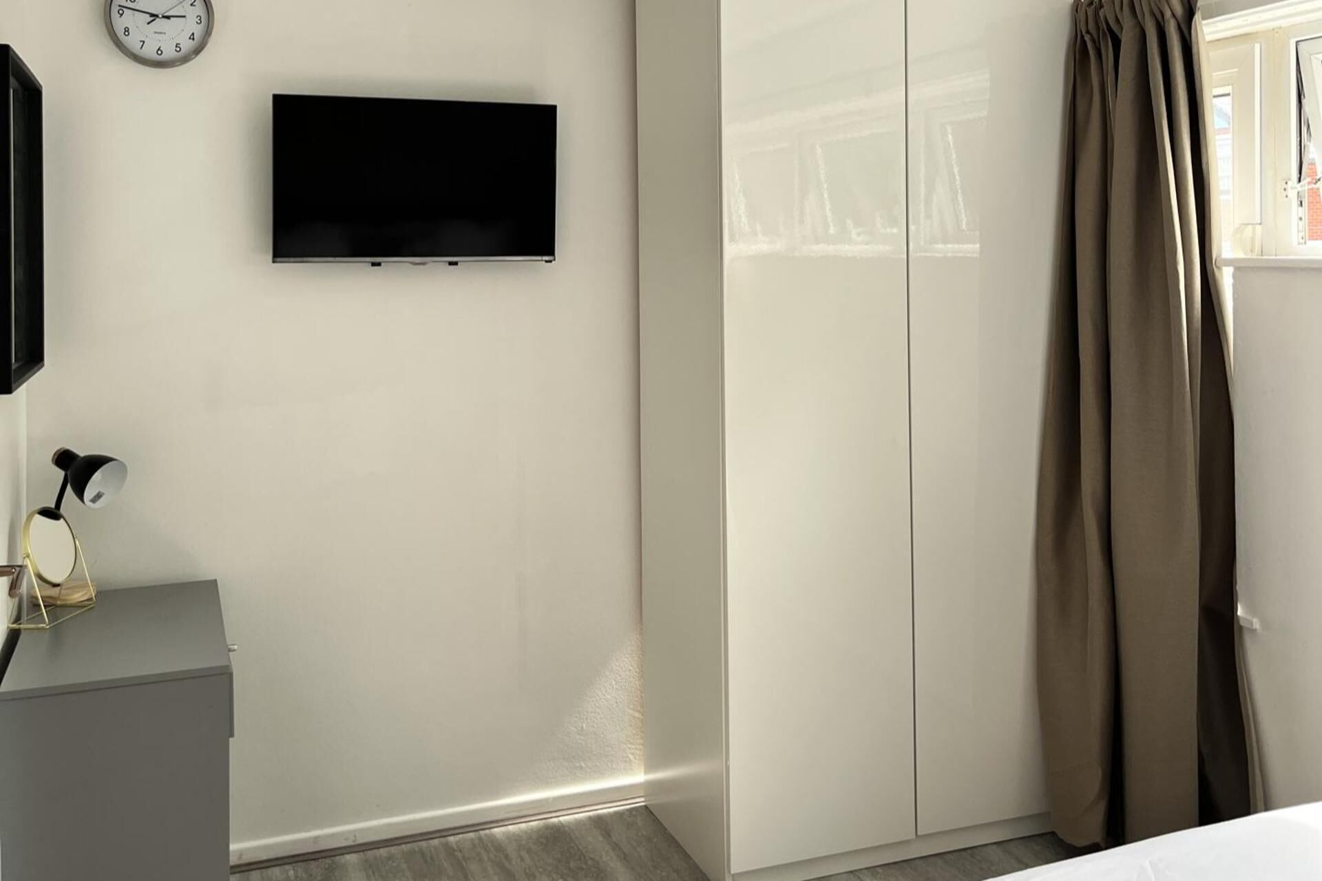 Un dormitorio con cama, armario, televisor montado en la pared y ventana con cortinas.