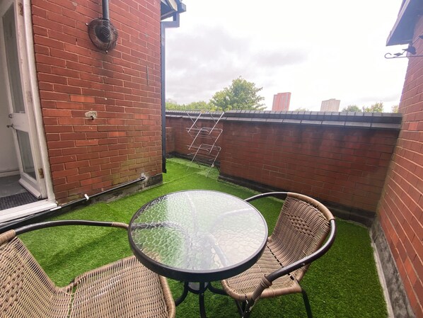 Un patio con una mesa de vidrio, sillas de mimbre, un muro de ladrillo y un tendedero.