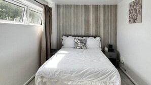 Una cama bien hecha con una colcha blanca y almohadas, una mesita de noche con una lámpara y una ventana con cortinas.
