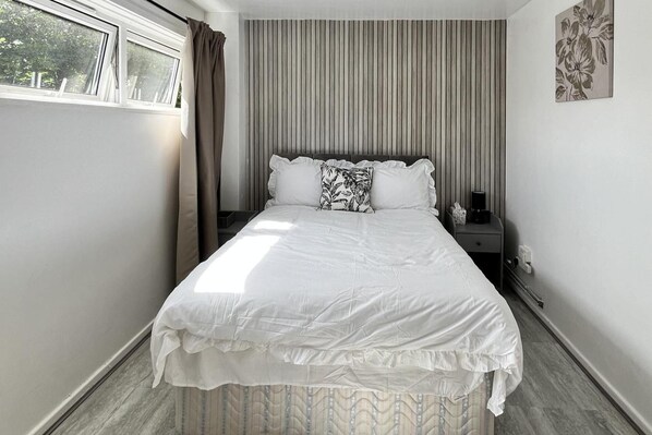A neatly made bed with a white comforter and pillows, a bedside table with a lamp, and a window with curtains. - Apartment - Sleeps 2 & Free Parking (Birmingham)