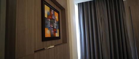A room with a framed abstract painting on the wall, a remote control on a shelf, and a window with dark curtains.