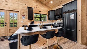 Ein Holzblockhaus-Innenraum mit Küchenbereich, der schwarzen Schränken, einem Edelstahl-Kühlschrank und einer Bar mit Barhocker-Bestuhung zeigt.