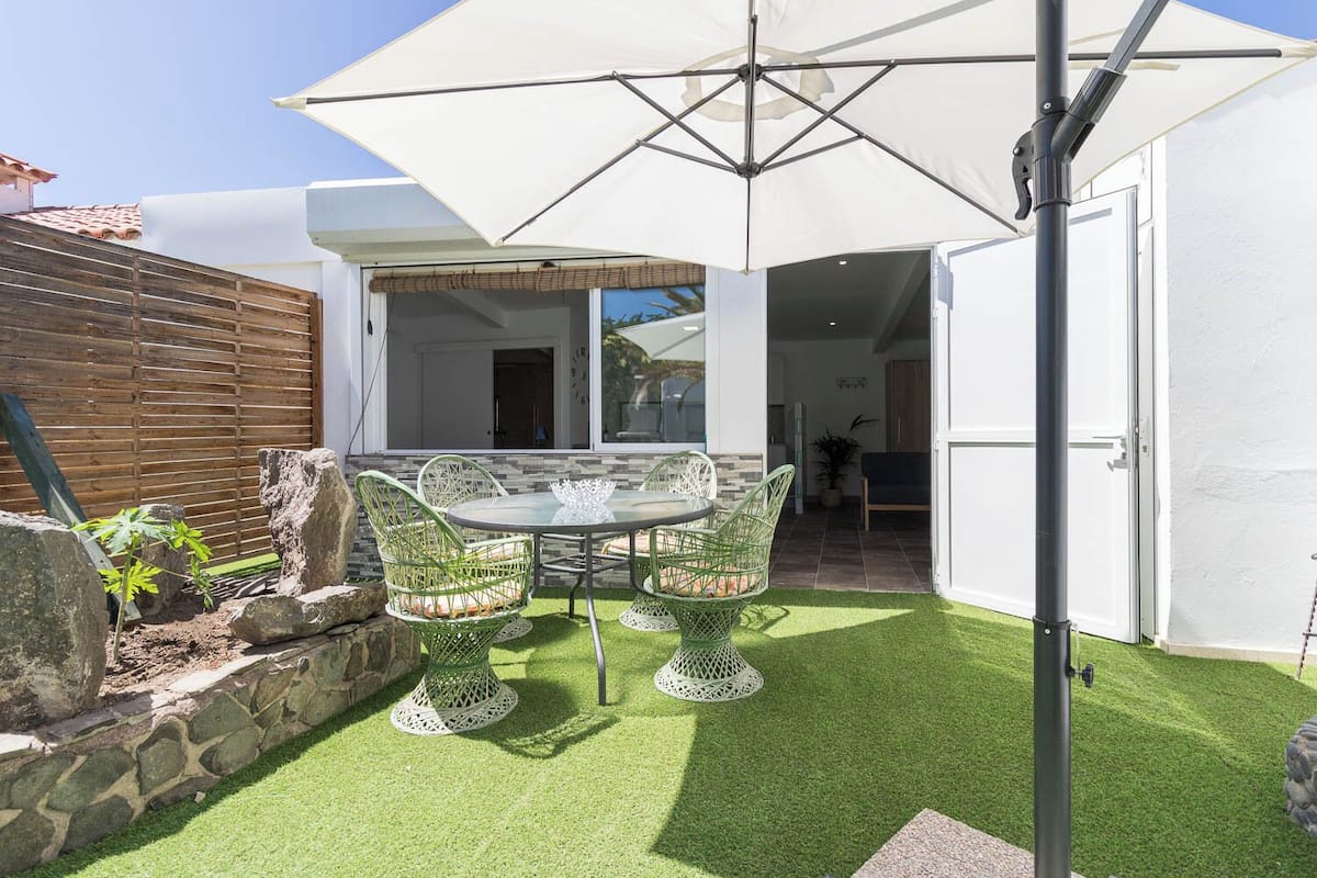 Um pátio com mesa e cadeiras, um guarda-sol branco e um muro de pedra.