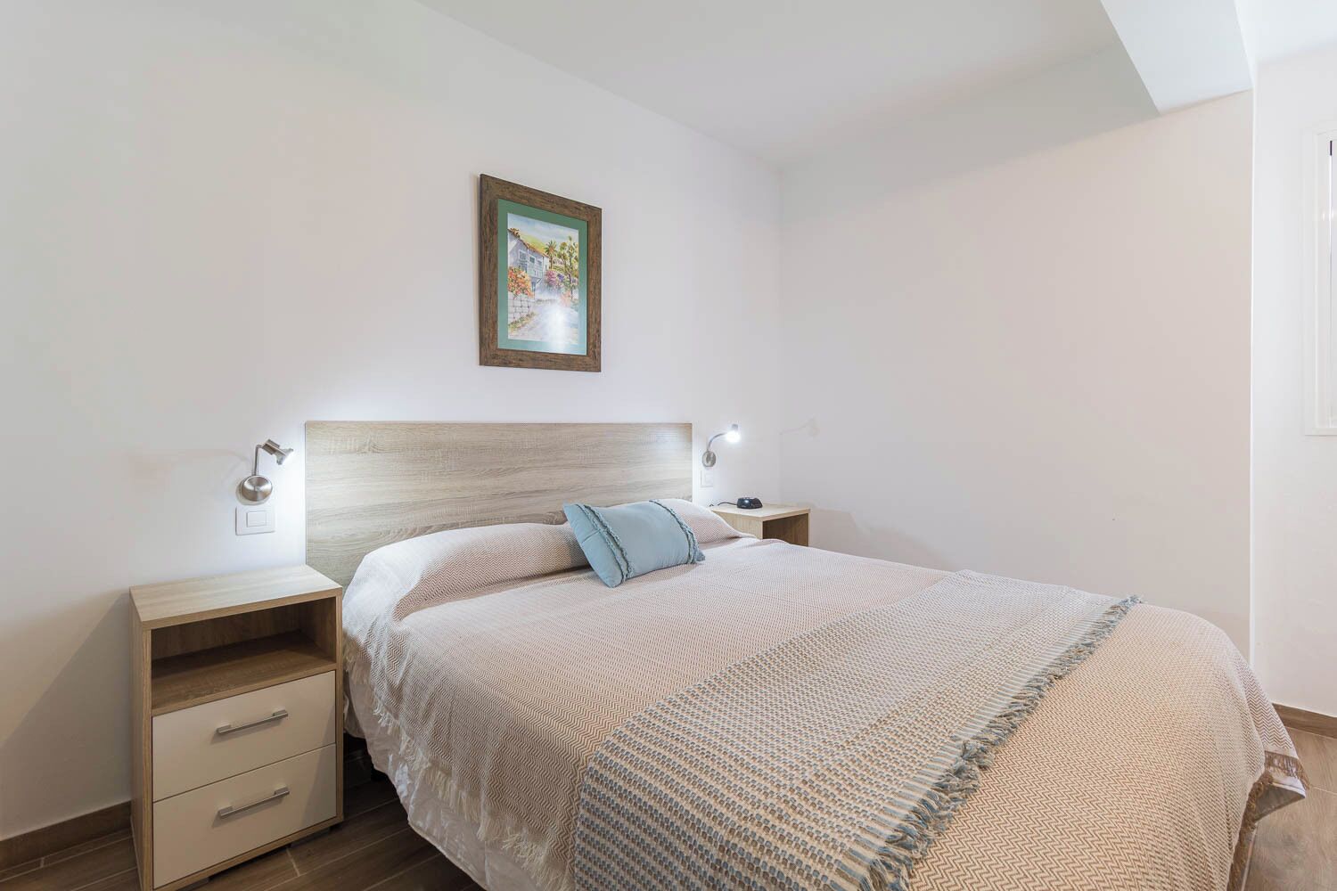 Ein Schlafzimmer mit einem hölzernen Kopfteil, einem Bett mit einer gemusterten Decke, Nachttischen mit Lampen und einem gerahmten Bild an der Wand.