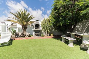 A well-maintained outdoor area with artificial grass, a stone barbecue, a white lounge chair, and a stone bench. - Bungalows Lapahi (San Bartolomé de Tirajana)