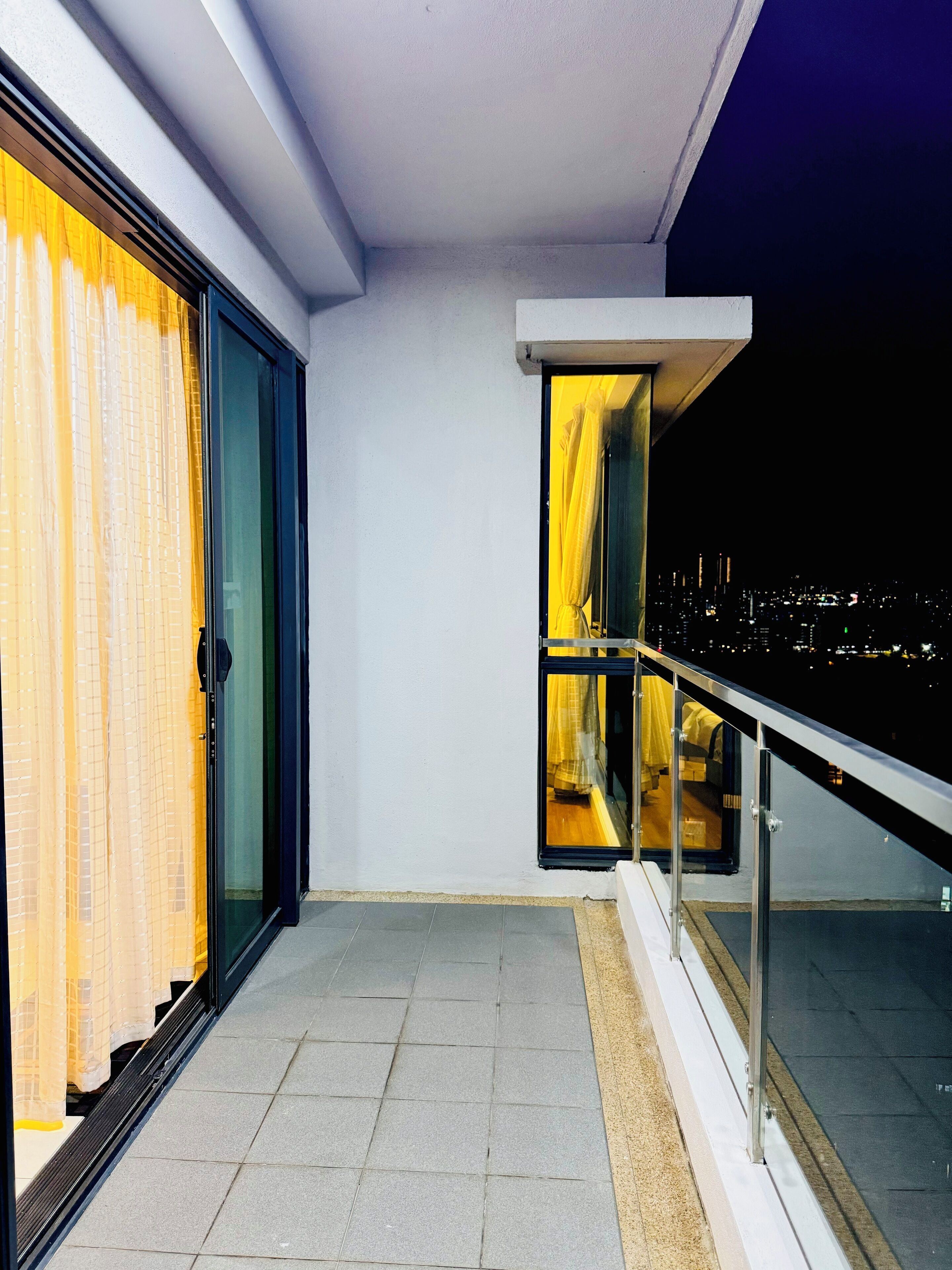 Un balcón con puerta de cristal, vista nocturna de la ciudad y barandilla.