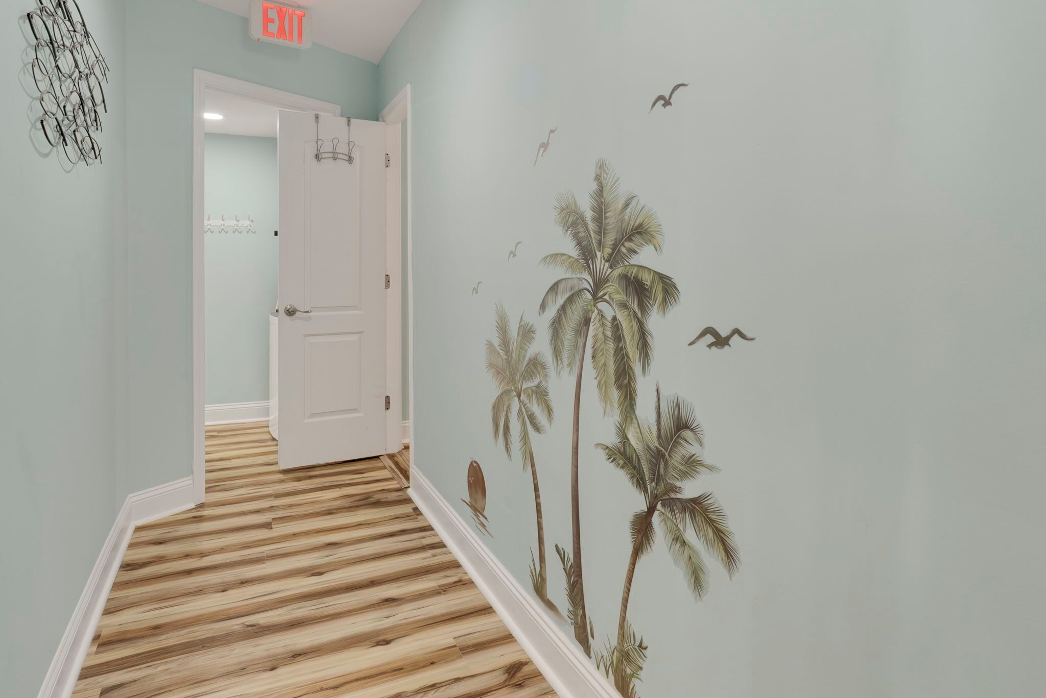 Un pasillo con piso de madera, una puerta con letrero de salida y una pared con un mural tropical.