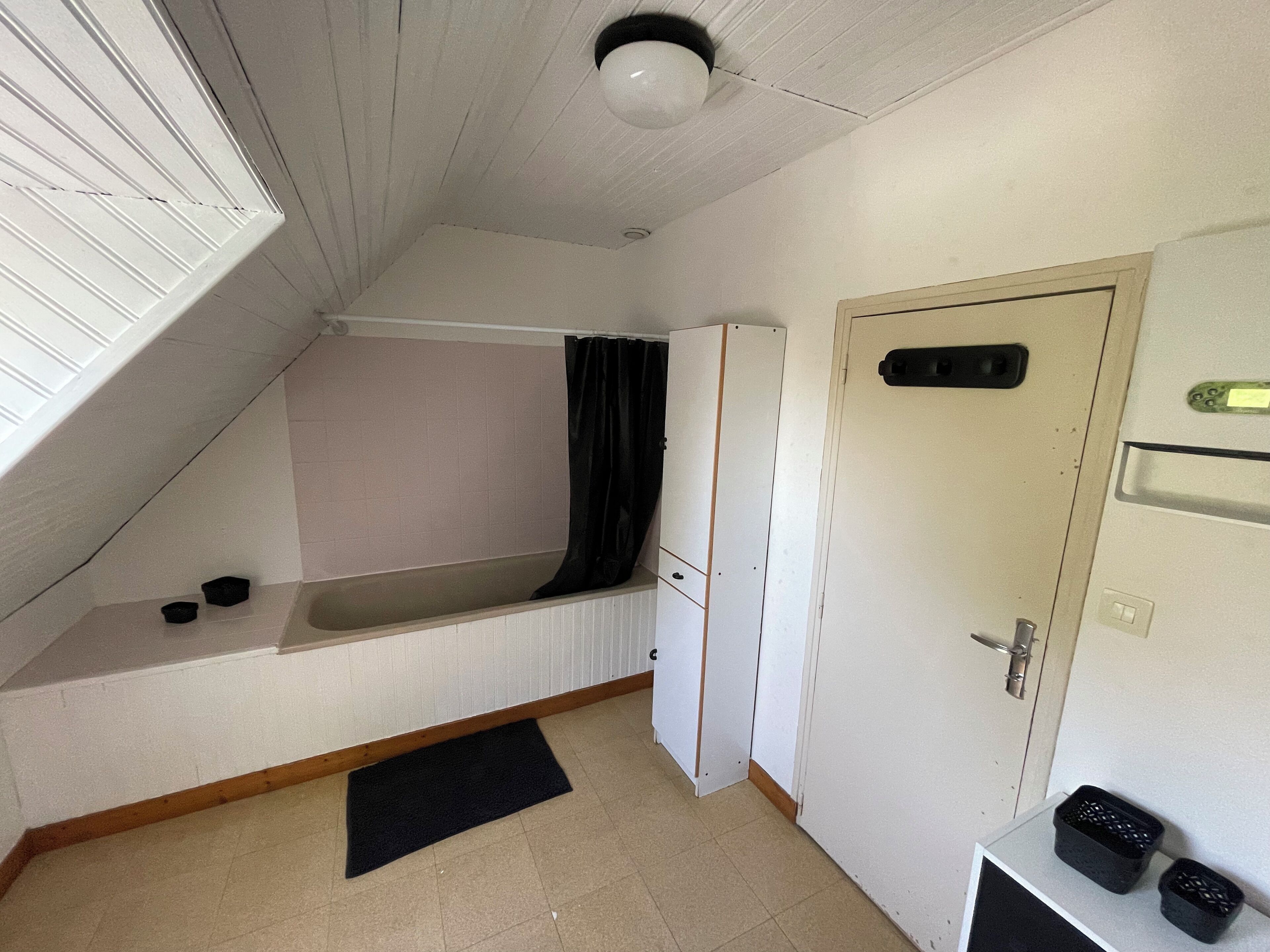 Une petite pièce aux dimensions réduites, dotée d’un plafond en pente, d’une baignoire intégrée, d’une porte avec une poignée et d’un petit tapis noir sur le sol.