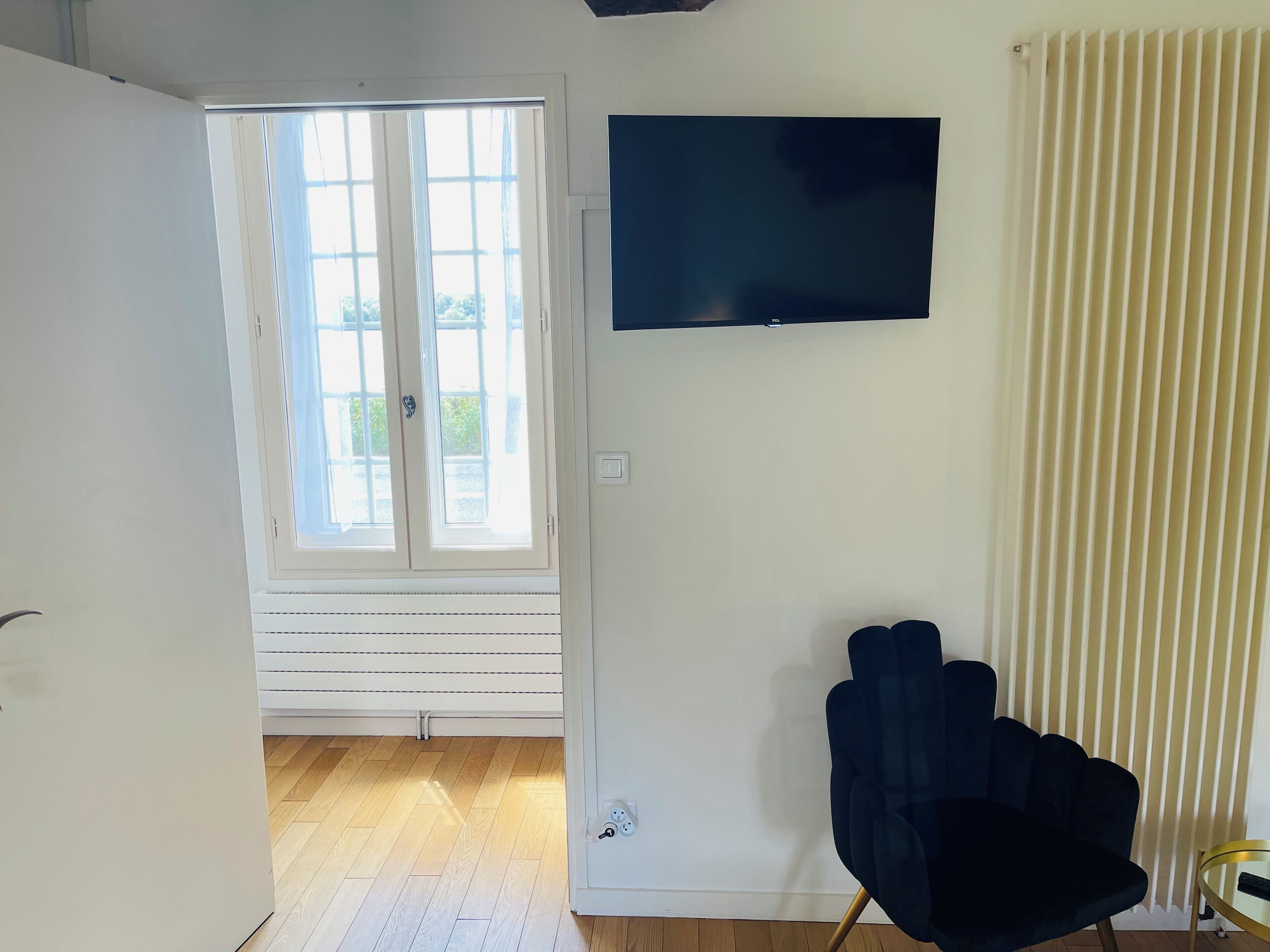 Ein Zimmer mit großem Fenster, einem flachbildfernseher an der Wand, einem dunklen Sessel und einem kleinen runden Tisch.