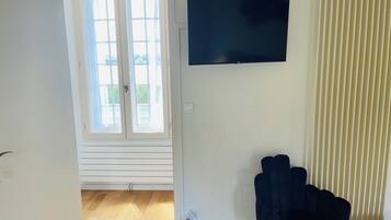 Ein Zimmer mit großem Fenster, einem flachbildfernseher an der Wand, einem dunklen Sessel und einem kleinen runden Tisch.