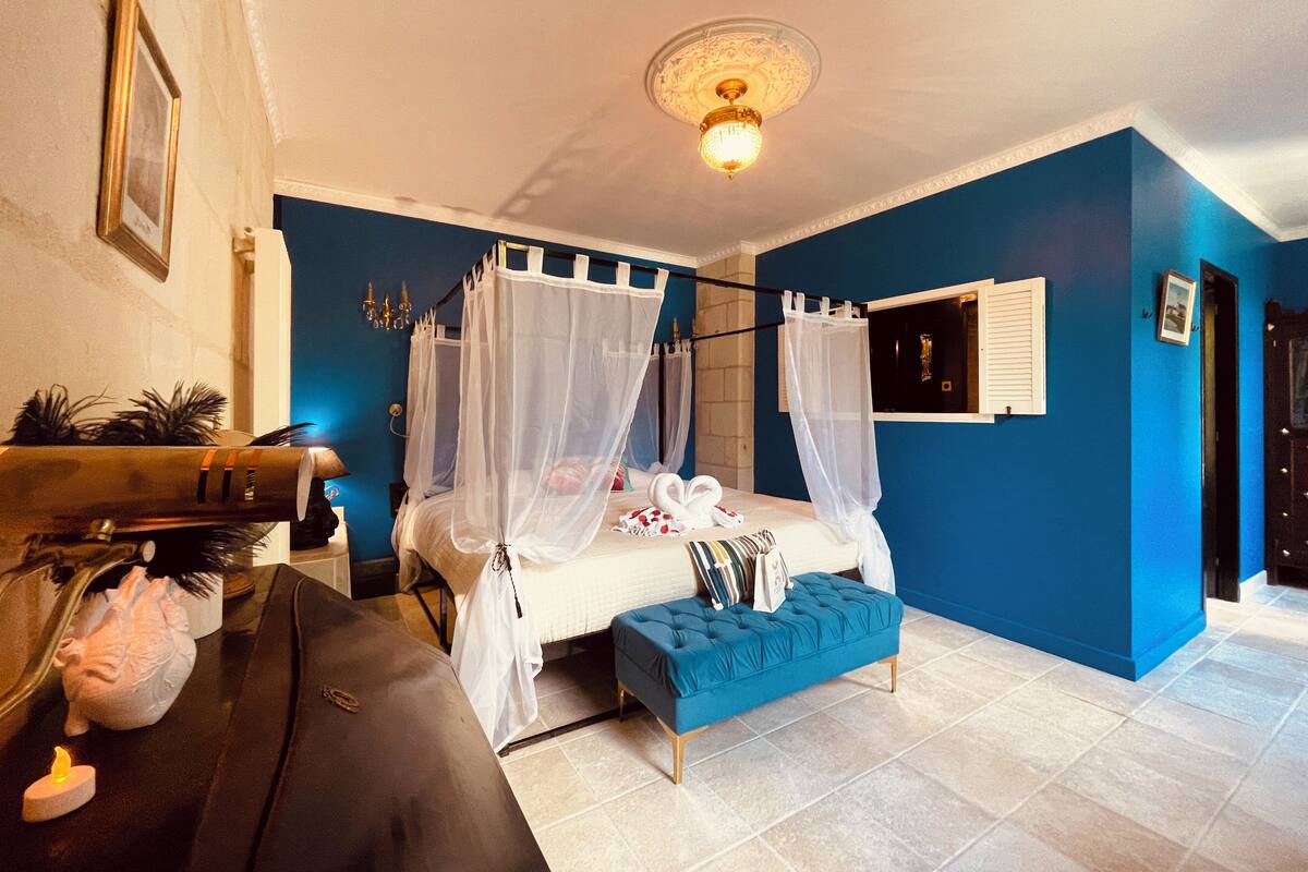 Quarto com cama, um banco azul, mesa de cabeceira, televisão na parede e luminária no teto.