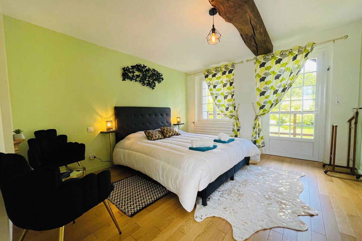 Quarto com uma cama grande, uma cadeira preta, piso de madeira e teto com vigas aparentes.