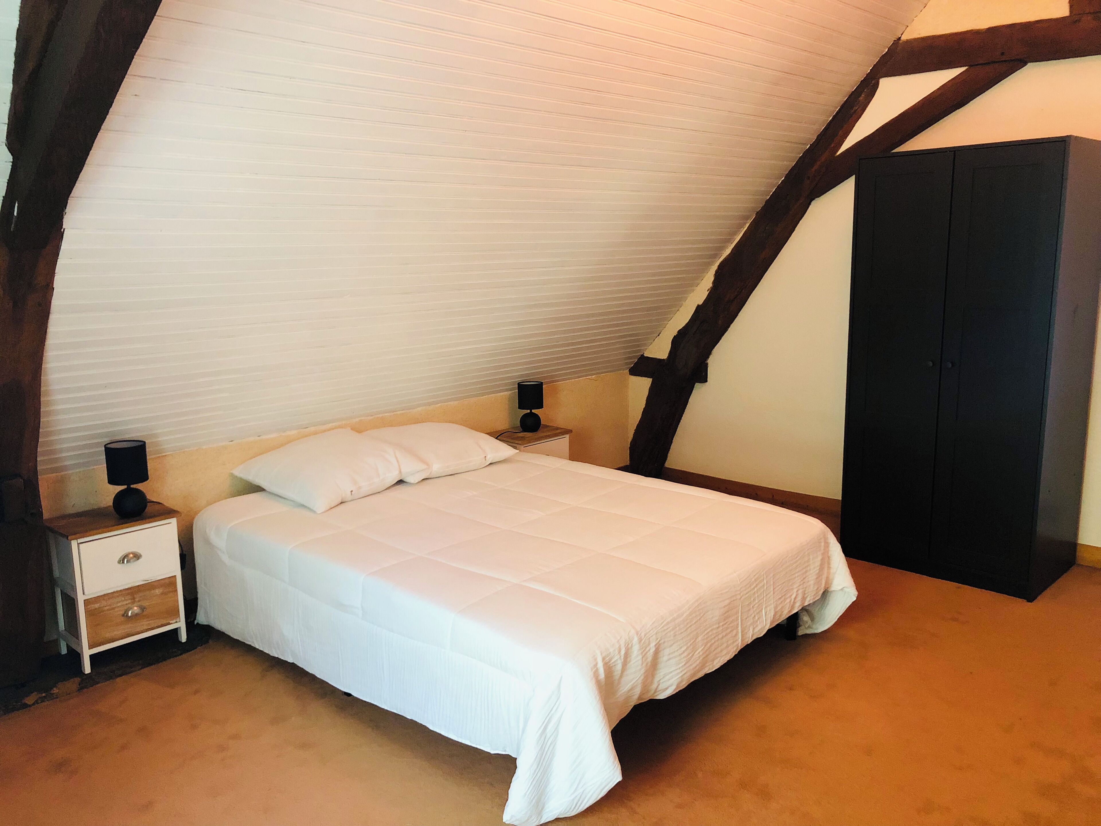 A bedroom with a sloped ceiling, a bed with white bedding, two bedside tables with lamps, and a dark wardrobe.