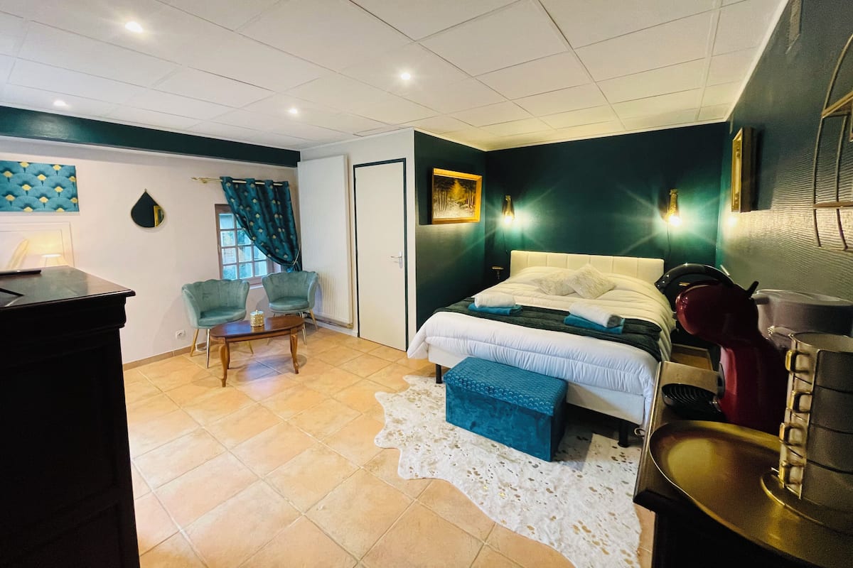 Quarto com cama, escrivaninha, cadeira, mesinha e janela com cortinas.