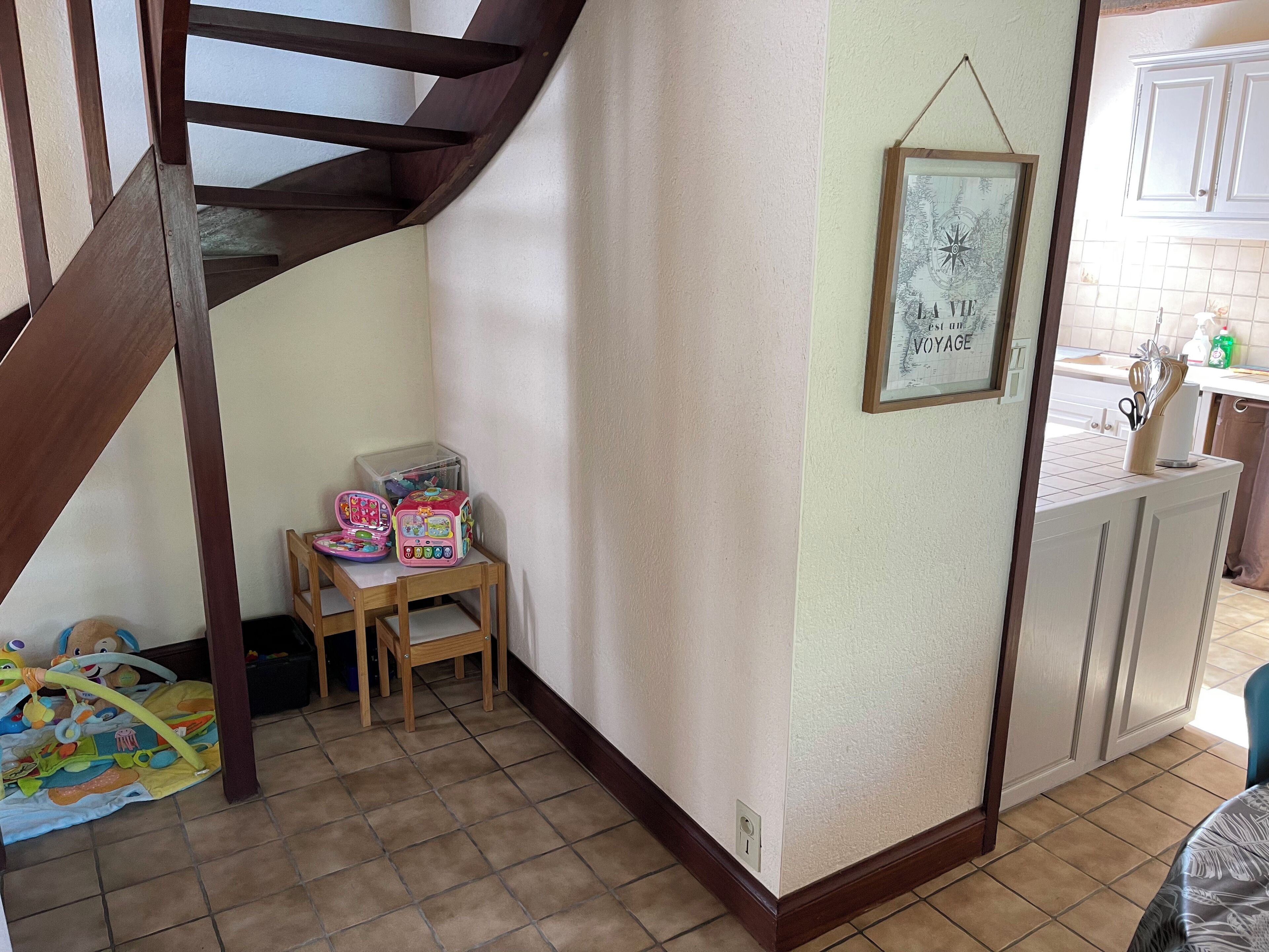 Un rincón de una habitación con una escalera de madera, una mesita con juguetes, un cuadro en la pared y una zona de cocina al fondo.