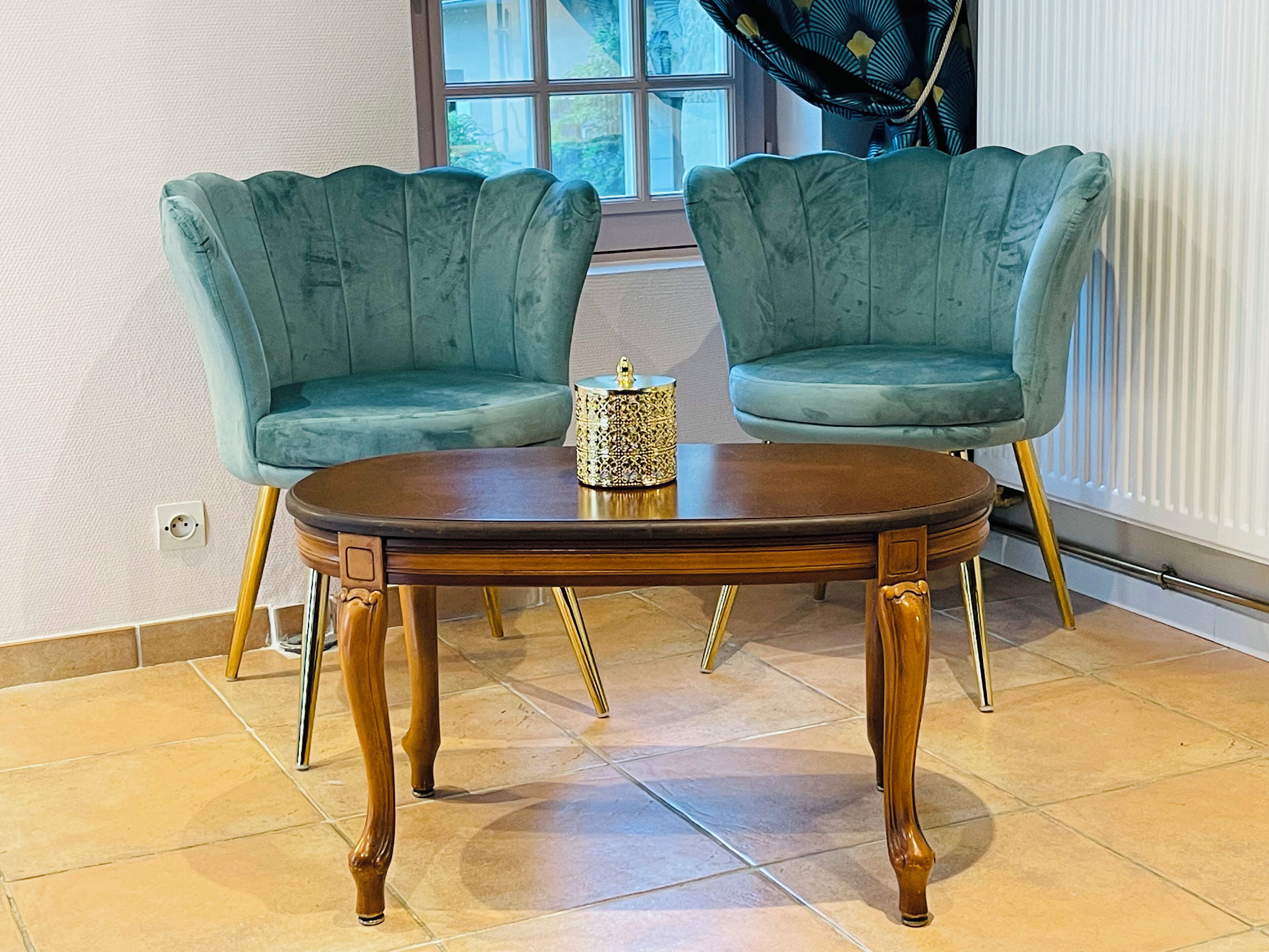 Dos sillones turquesa con patas doradas alrededor de una mesa redonda de madera con un centro decorativo.