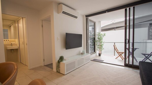Una sala de estar moderna con un televisor de pantalla plana, un ventanal corredero que da a un balcón y una pequeña planta en maceta.