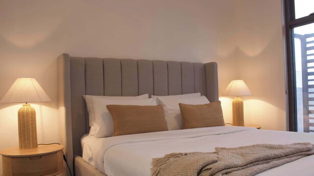 A neatly made bed with a grey upholstered headboard, two bedside lamps, and a window with blinds. - 2BR with Parking | Walk to QPAC & South Bank (South Brisbane)
