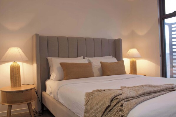 A neatly made bed with a grey upholstered headboard, two bedside lamps, and a window with blinds.